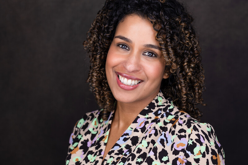 Woman with curly dark hair and bright smile wearing a colorful patterned blouse against a dark background.