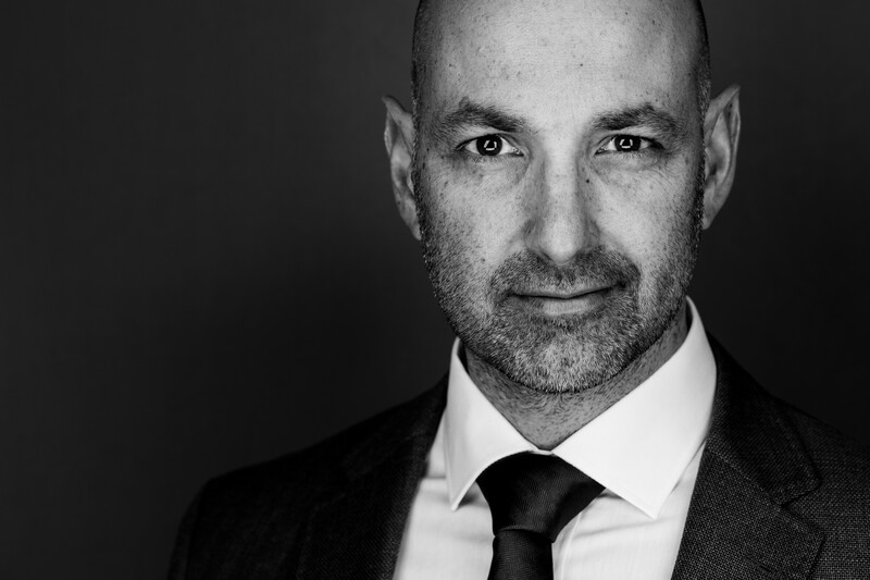 Professional headshot of a bald man with a beard wearing a dark suit and black tie against a dark background.