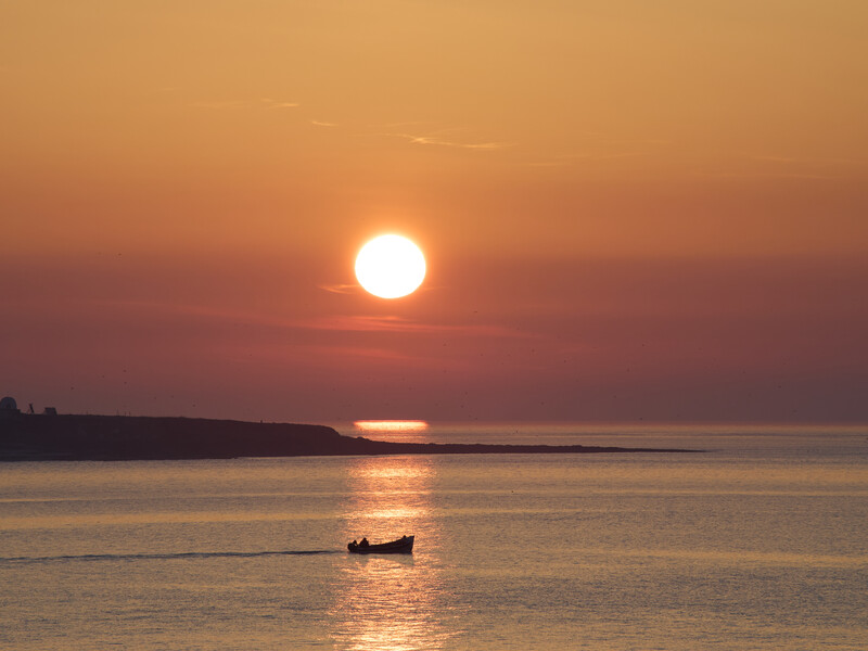 Silhouetted boat on calm water during golden-hour sunset with large sun reflected on the sea.
