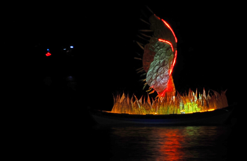 Nighttime long exposure photograph of a sculptural art installation with illuminated stone and spiky orange-yellow elements reflected in still water.