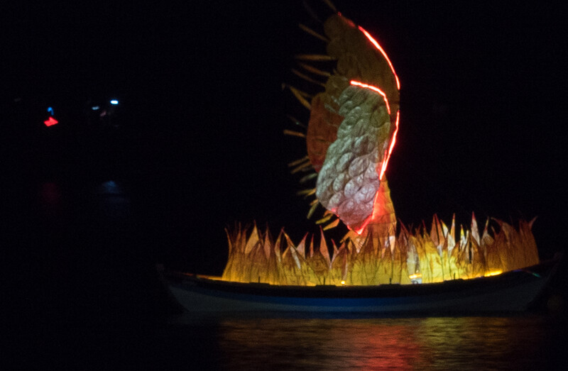 Illuminated sculptural installation of a large fish head with open mouth and gill spines, glowing with warm orange and yellow LED lighting against dark water.