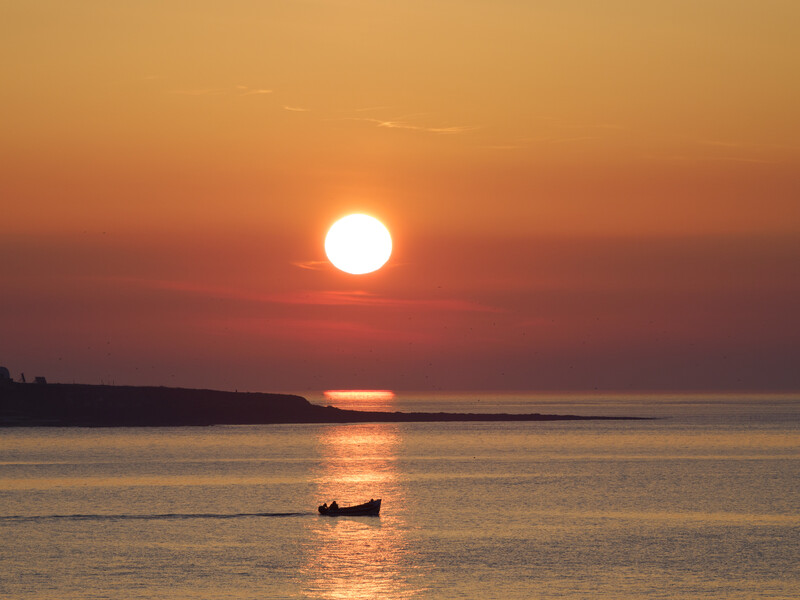 Silhouetted fishing boat on calm water with large sun setting on the horizon beneath an orange and purple sky.