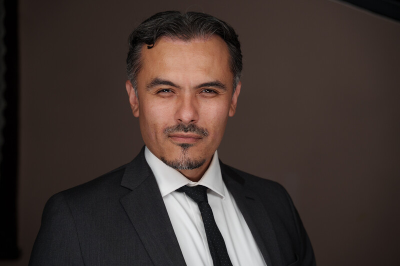 Professional headshot of a man in a dark suit and black tie against a neutral brown background.