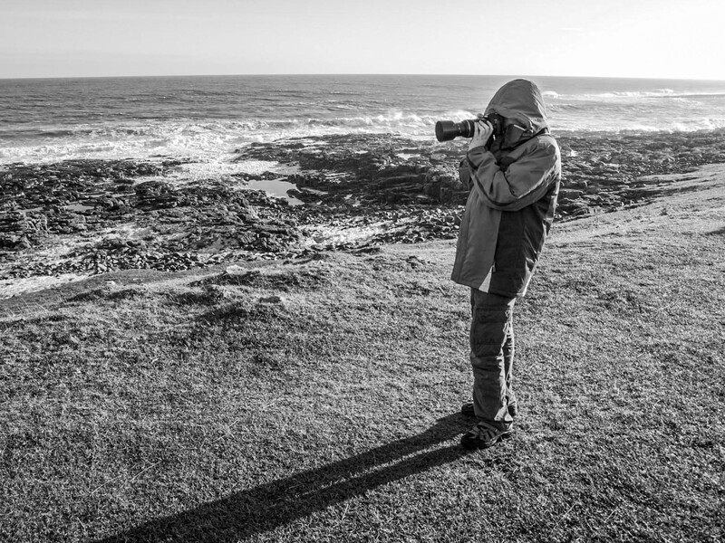 Photographer standing on a rocky beach, holding a telephoto lens to their eye while framing a seascape shot.