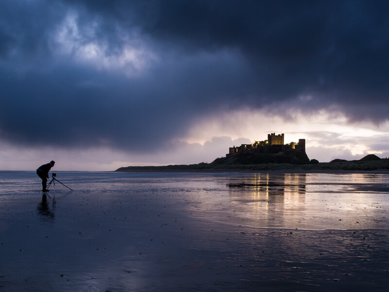 Person wading in shallow water on a beach at dusk, with a castle silhouetted on a distant tidal island beneath stormy skies.