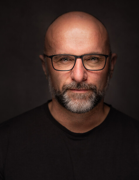 Man with glasses and beard wearing dark shirt against black background.