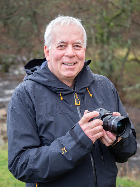 Older man with white hair smiling while holding a compact digital camera outdoors.