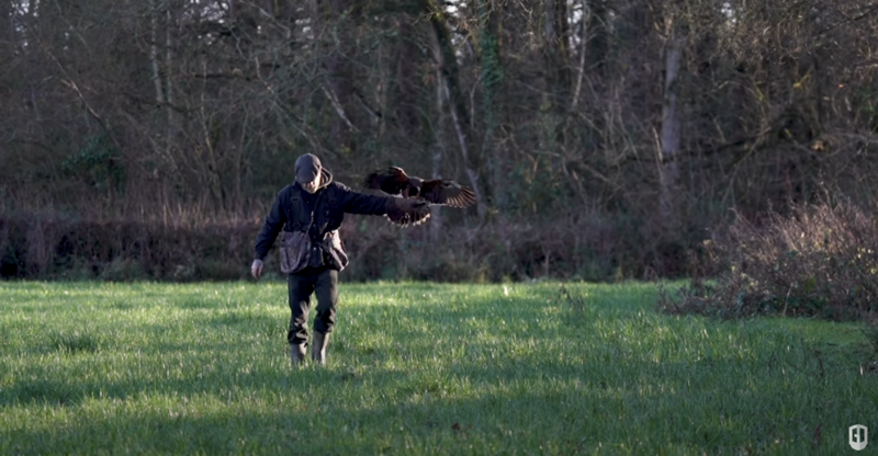 Person in dark blazer performing dynamic pose with extended arm in open field with bare trees in background.
