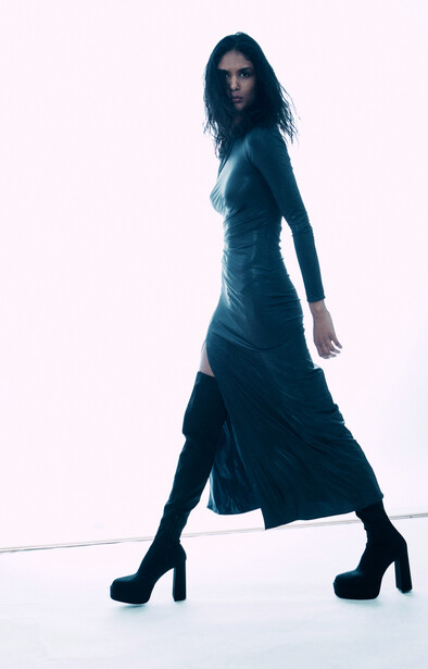 Woman in dark draped dress and platform heels posing against white background.