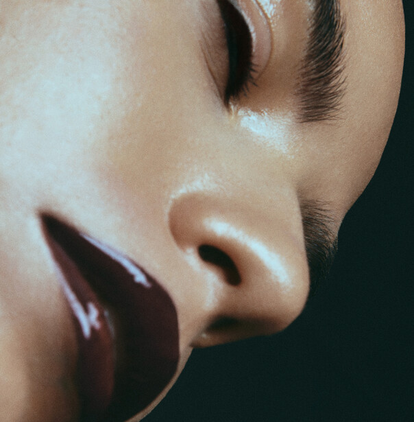 Close-up profile of a face with glossy lips and defined eyebrow against dark background.
