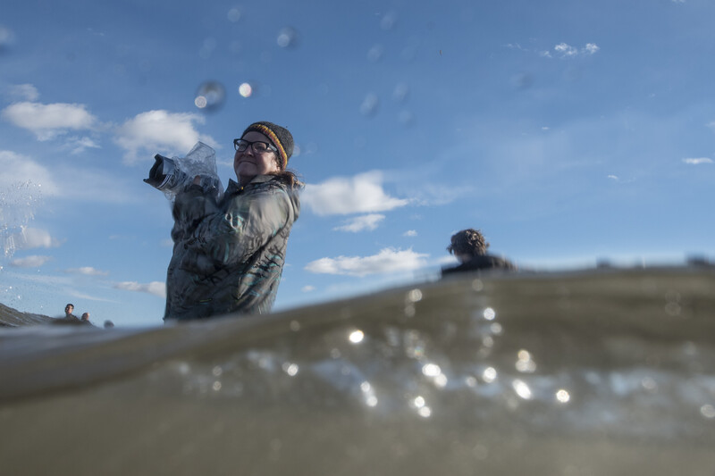 Photographer holding camera above water surface with bokeh water droplets in foreground and landscape in background.