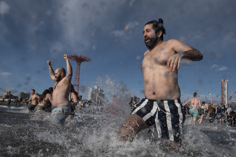 Shirtless men running through shallow ocean water with splashing waves on a sunny day.