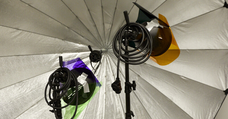 Studio lighting setup with three monolight heads mounted on stands inside a large white umbrella reflector.