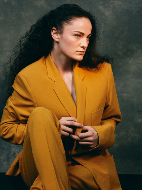 Woman in mustard yellow blazer against muted green background.