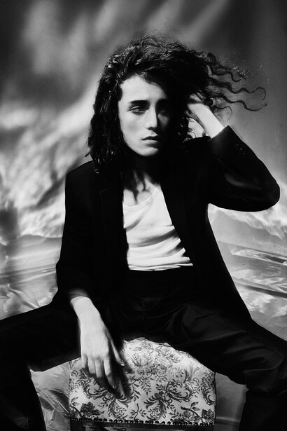 Portrait of a person with long dark hair in a black blazer, posed against a blurred background.