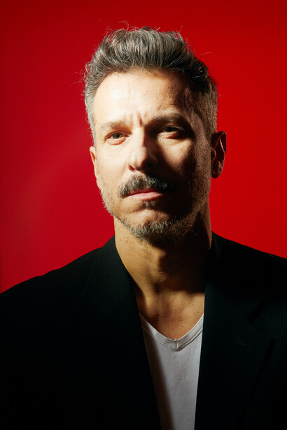 Man with gray hair and beard against solid red background, wearing black blazer over light gray shirt.