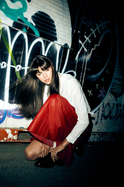 Woman in red dress and white jacket crouching against graffitied wall at night.