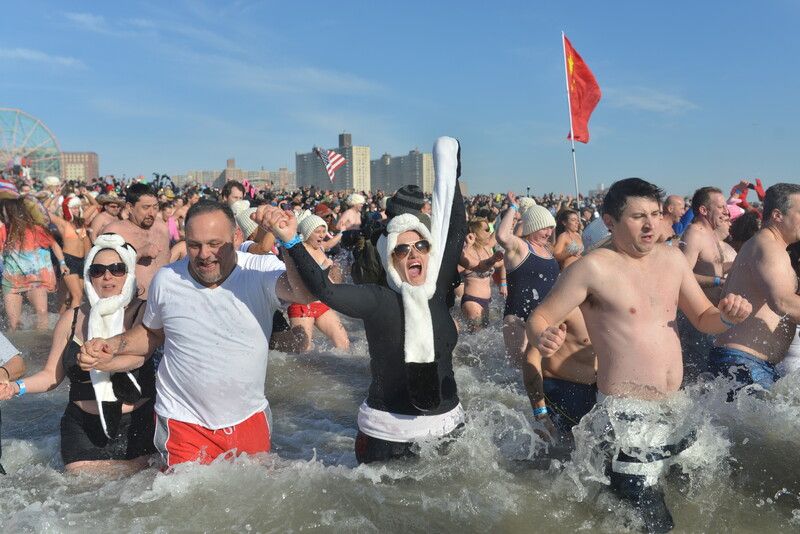 Large crowd wading in ocean water with city skyline and red flag visible in background.