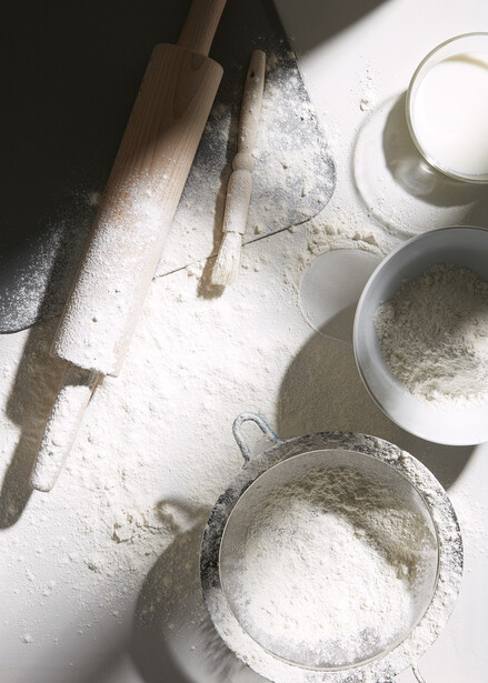 Overhead arrangement of baking ingredients including flour in bowls and sieves, a wooden rolling pin, and a wooden spoon on a white surface.
