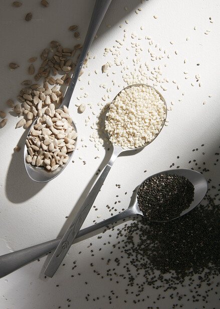 Three spoons holding different seeds and grains on white surface with scattered particles and shadows.