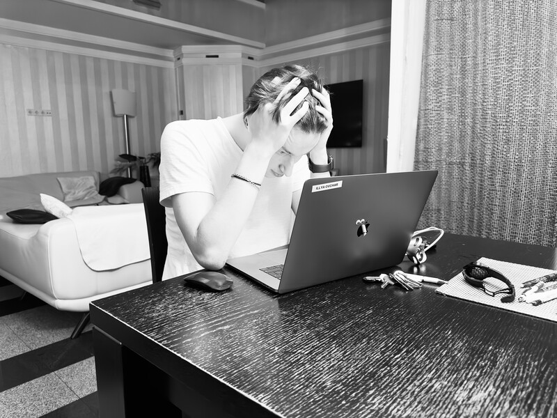Man in hotel room experiencing stress while working on laptop at desk.