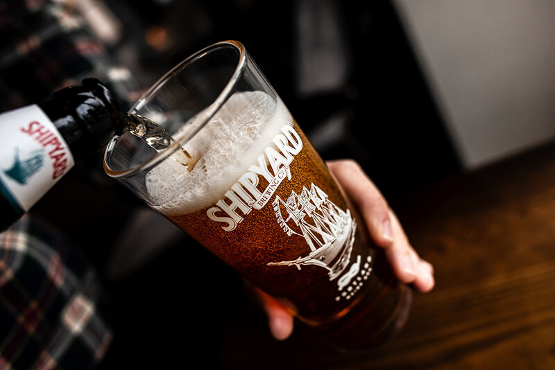 Hand holding a glass of beer with foam head and condensation, showing amber liquid inside.