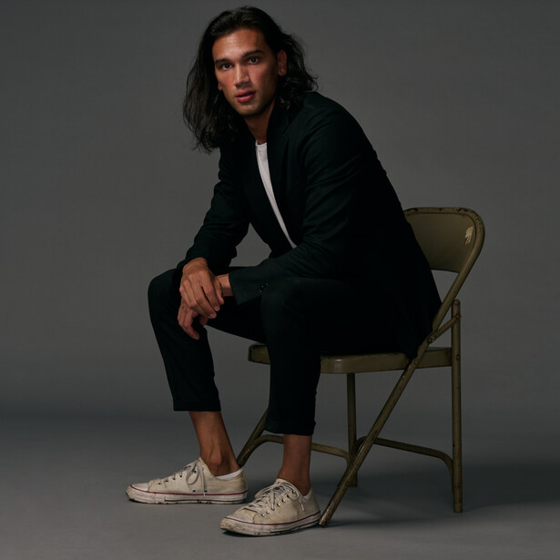 Person in black blazer and pants sitting on a folding chair against gray background.