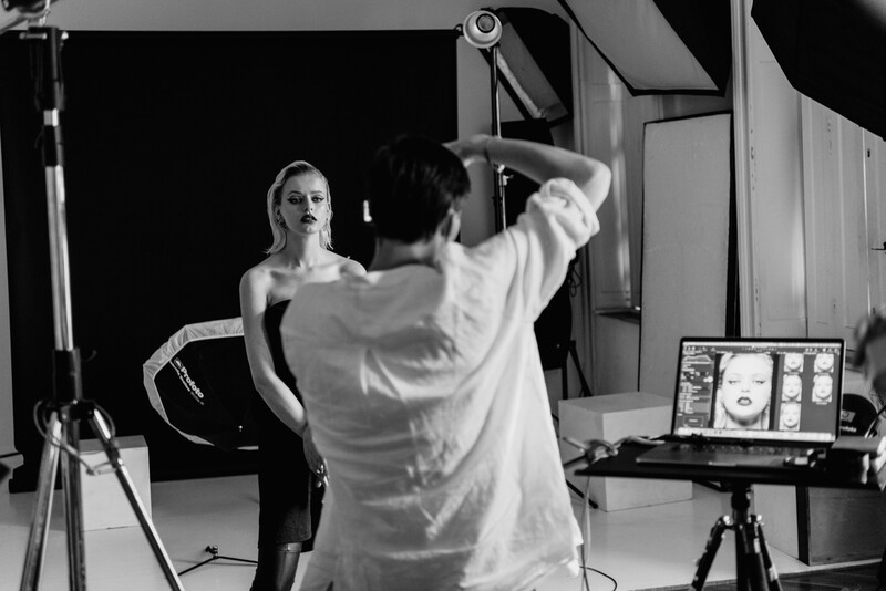 Photographer adjusts studio lighting equipment while model sits on set during a black and white portrait shoot.