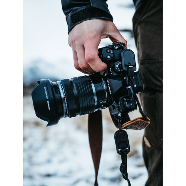 Photographer holding a mirrorless camera with telephoto lens attached, wearing dark jacket and camera strap.