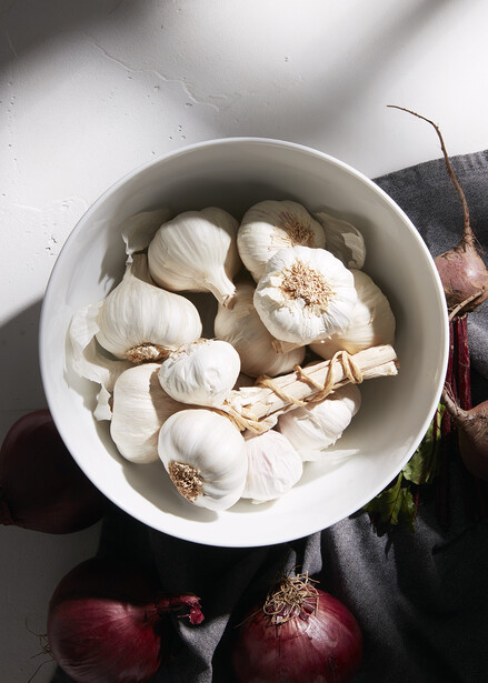 Overhead view of a white bowl containing garlic cloves and whole garlic bulbs with red onions beside it.