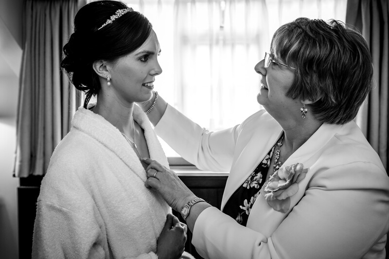 Woman in wedding dress and tiara smiles at an older woman adjusting her necklace in a black and white portrait.