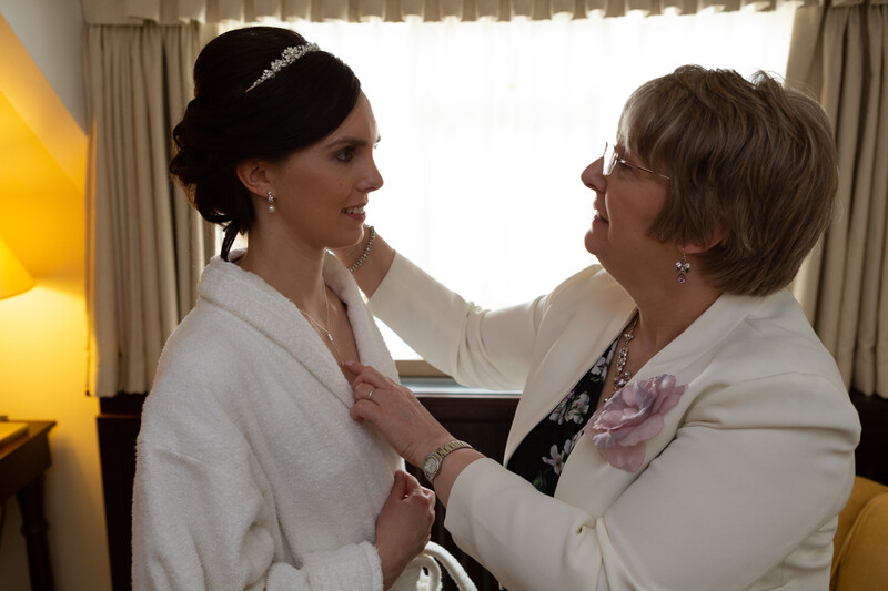 Woman in white cardigan and tiara being adjusted by older woman in cream blazer indoors.