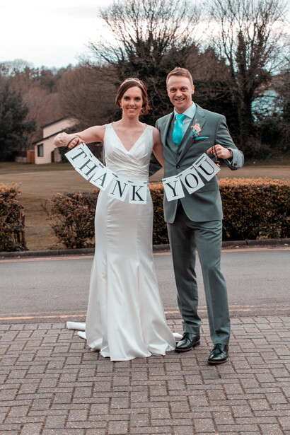 Bride and groom holding a banner reading "THANK YOU" while posing outdoors on a brick pathway.