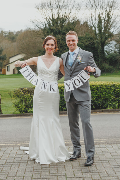 Bride and groom holding a banner with initials while standing on a brick driveway.