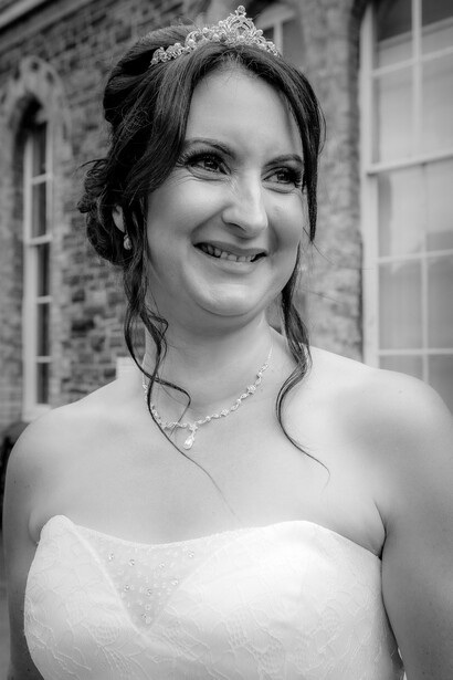 Bride in strapless white wedding dress with pearl necklace and upswept hairstyle, smiling outdoors against brick building.