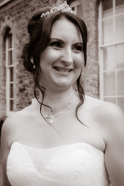 Bride in white strapless wedding dress wearing tiara and pearl necklace, smiling outdoors.