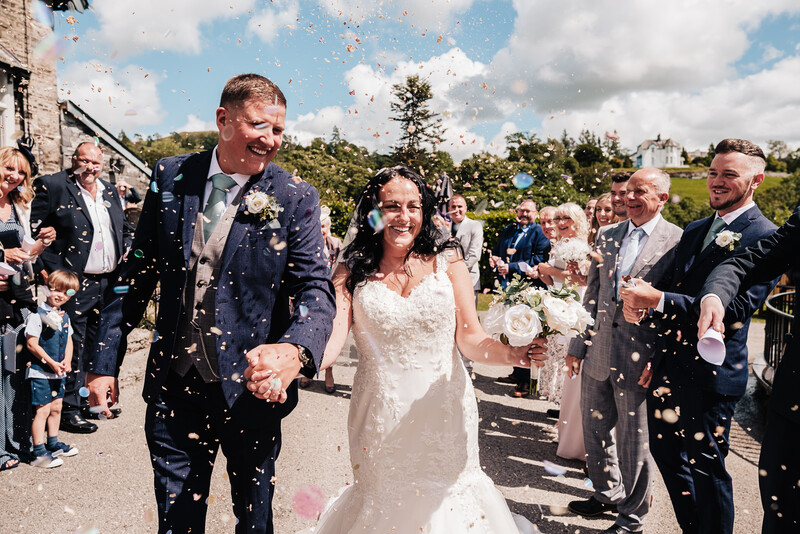 Newlyweds walk through a shower of confetti as wedding guests celebrate on a sunny day.
