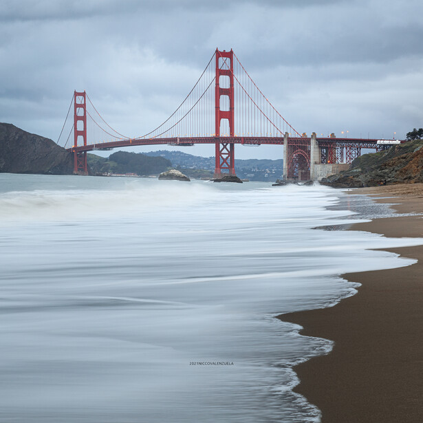 Golden Gate Bridge spanning across San Francisco Bay under cloudy skies, photographed from a sandy beach with soft waves in the foreground.