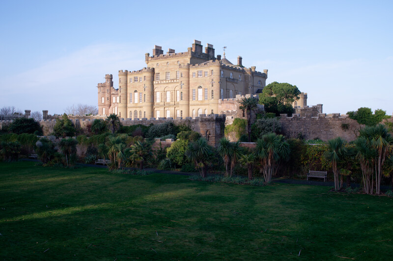 Golden-hued stone castle on elevated ground surrounded by manicured lawn and palm trees.