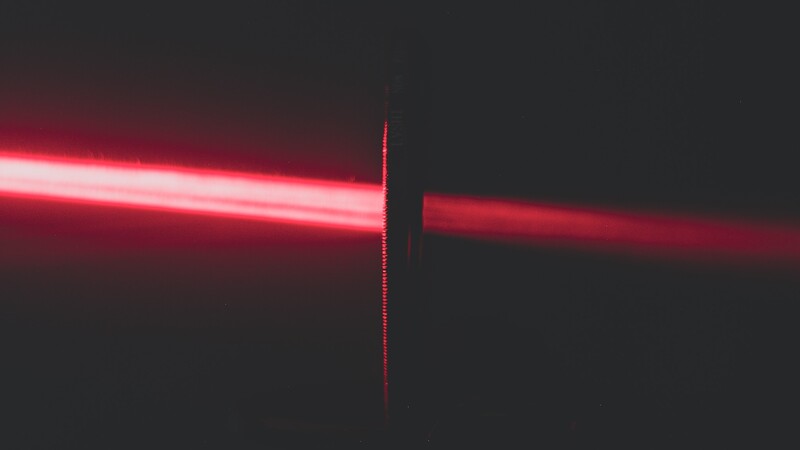 Red laser beam passing through a vertical black obstruction against a dark background.