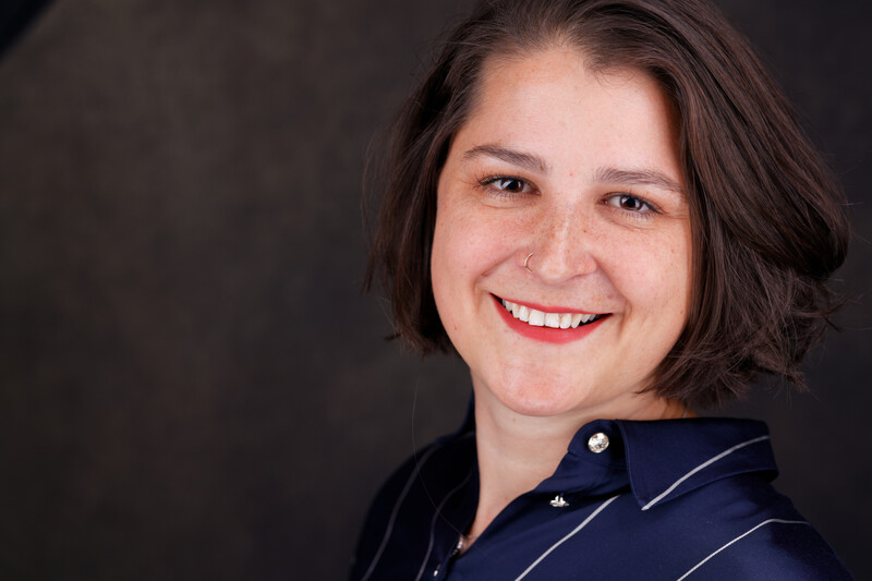 Professional headshot of a woman with shoulder-length dark hair wearing a dark blue shirt, smiling at the camera against a dark background.