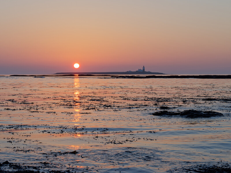 Sun setting over a calm bay with a distant lighthouse silhouetted on the horizon.