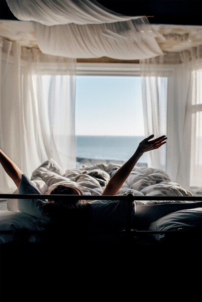 Person stretching in bed with ocean view through sheer curtains and open window.