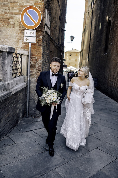 Bride and groom walking down a narrow European cobblestone street lined with historic stone buildings.