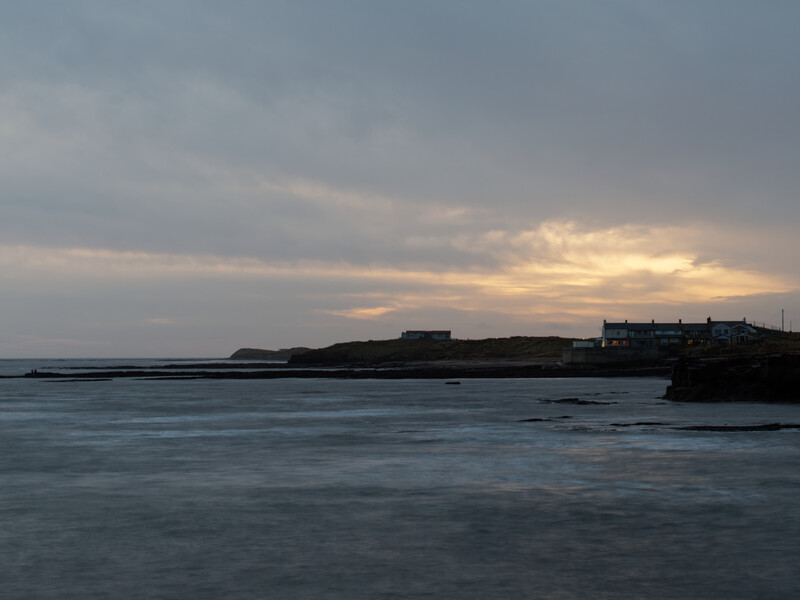 Coastal landscape at dusk with silhouetted buildings on a rocky headland under a dramatic cloudy sky.