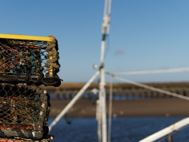 Weathered lobster trap in sharp focus with wind turbine and bridge blurred in background over water.