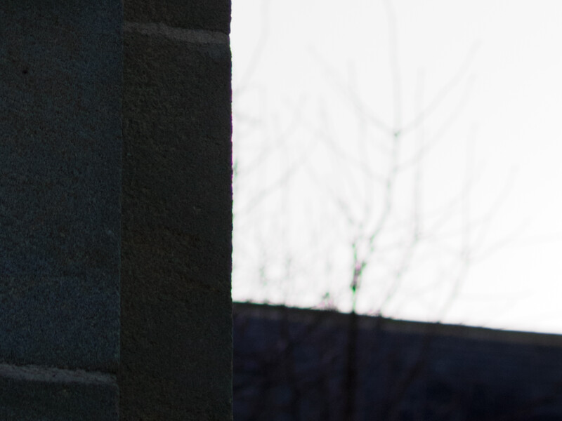 Dark architectural element with sharp geometric lines against a blurred overcast sky and bare branches.