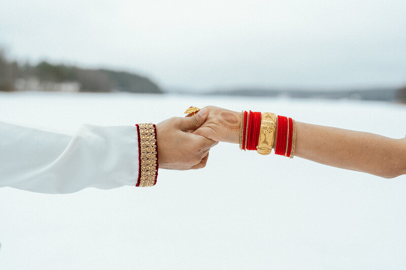 Two people holding hands outdoors, one wearing a white sweater and tan bracelet, the other wearing red and gold bangles, with a frozen lake and forested shoreline in soft focus behind them.