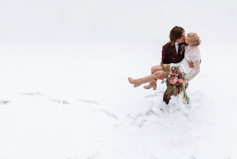 Groom carrying bride through deep snow while kissing, both in winter wedding attire.