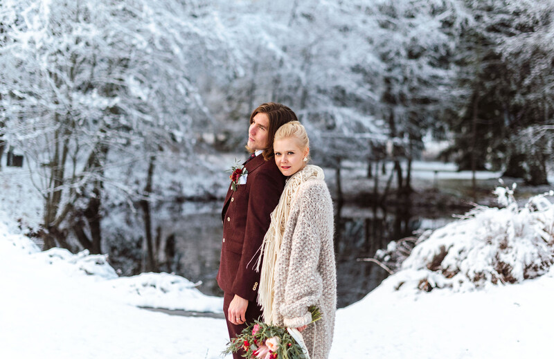 Two people in winter clothing standing in a snowy forest landscape with evergreen trees.